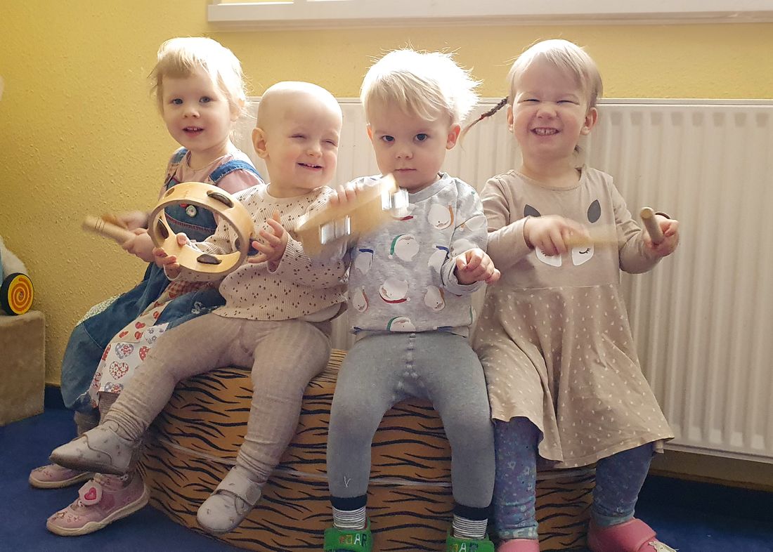 Vier Kleinkinder sitzen nebeneinander und musizieren fröhlich mit Tamburin und Klanghölzern